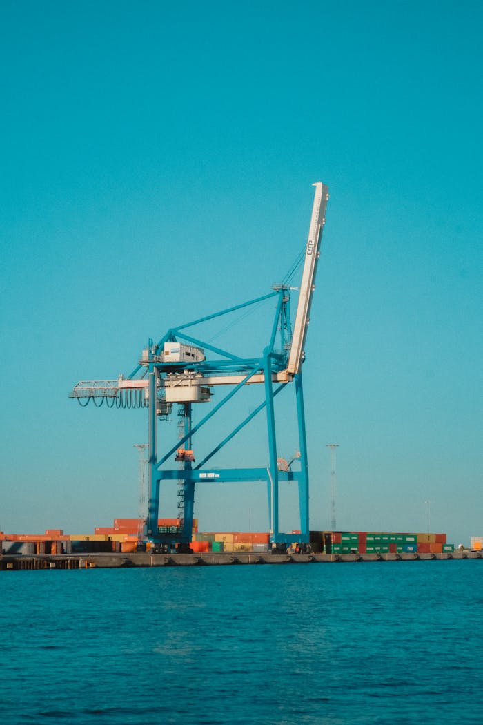 hero-img-01 A large blue cargo crane at a shipping port with containers on a clear day.